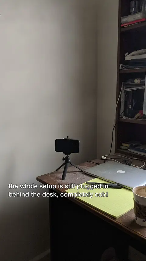A phone on a tripod in the corner of an unlit room, captioned "the whole setup is still plugged in behind the desk, completely cold".